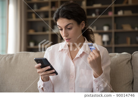 Woman holds debit card staring at smartphone screen Woman holds debit card staring at smartphone screen 90014746