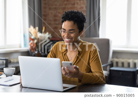 African woman sit at desk holds cellphone staring at laptop 90015005