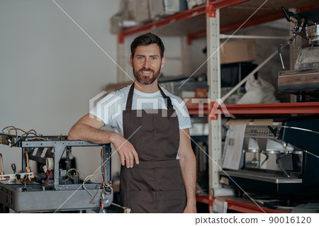 Portrait of repairman in uniform standing on workshop of coffee machines warehouse Portrait of repairman in uniform standing on workshop of coffee machines warehouse 90016120