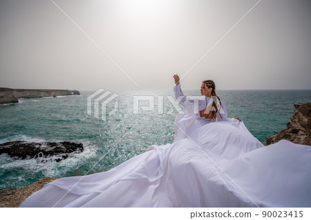 Happy freedom woman on the beach enjoying and posing in white dress. Rear view of a girl in a fluttering white dress in the wind. Holidays, holidays at sea. 90023415