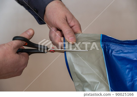 Hands with scissors cut a gray-blue bag. A middle-aged man opens a bag of pet food. Side view. Inside the room. Selective focus. Hands with scissors cut a gray-blue bag. A middle-aged man opens a bag of pet food. Side view. Inside the room. Selective focus. 90025166