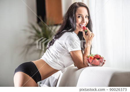 Woman lenaing against the couch is seductively looking into the camera taking a strawberry into her mouth 90026373