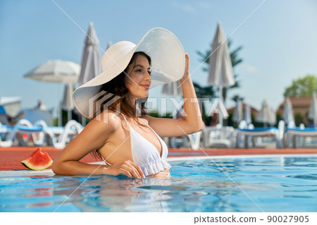 Side view of brunette young female in white swimsuit touching hat. Seductive girl standing in water in pool, looking forward, smiling, sunbathing. Concept of vacation and youth. Side view of brunette young female in white swimsuit touching hat. Seductive girl standing in water in pool, looking forward, smiling, sunbathing. Concept of vacation and youth. 90027905