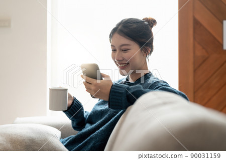 Cheerful young asian woman using mobile phone while sitting on a couch at home. Cheerful young asian woman using mobile phone while sitting on a couch at home. 90031159