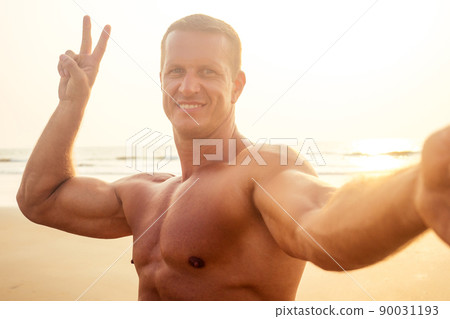 handsome successful male athlete fitness model photographing selfie portrait on phone beach paradise sunset. man on vacation in the tropic talking on the smartphone online with friends and family handsome successful male athlete fitness model photographing selfie portrait on phone beach paradise sunset. man on vacation in the tropic talking on the smartphone online with friends and family 90031193