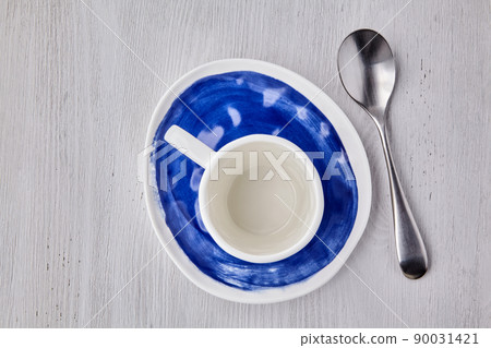Empty white cup for hot drinks with a spoon on a blue plate on a white wooden table Empty white cup for hot drinks with a spoon on a blue plate on a white wooden table 90031421