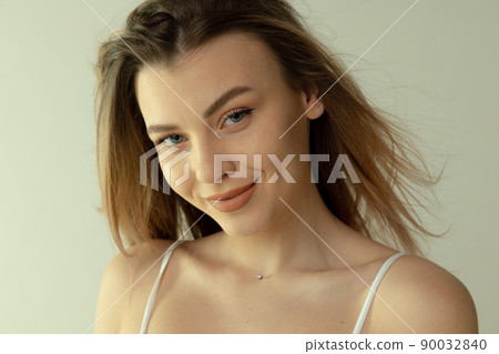 Close-up portrait of beautiful young blonde girl with natural makeup posing isolated over grey studio background. Tenderly smiling 90032840