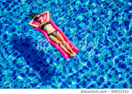 copy space spf and sunscreen beautiful brunette girl floating in the pool water.woman swimming and relaxing on pink inflatable mattress in blue swimmingpool remote work and freelance top view 90032932