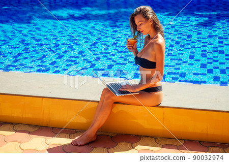 Young female freelancer working on laptop near the pool . Busy woman at holidays. Distant remote work concept. Copy space girl in black bikini beach holding a glass with a non-alcoholic fruit cocktail Young female freelancer working on laptop near the pool . Busy woman at holidays. Distant remote work concept. Copy space girl in black bikini beach holding a glass with a non-alcoholic fruit cocktail 90032974
