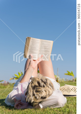 Woman lying and reading her favorite book on a meadow covered with fresh green grass on a sunny summer or spring day. Woman lying and reading her favorite book on a meadow covered with fresh green grass on a sunny summer or spring day. 90034728