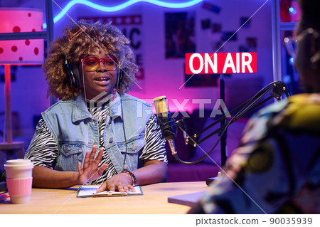 Stylish young African American woman sitting at desk in front of male co-worker discussing something during podcast or broadcast recording Stylish young African American woman sitting at desk in front of male co-worker discussing something during podcast or broadcast recording 90035939