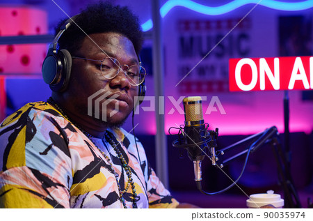 Medium close-up of self-confident young gen Z African American man wearing headphones doing broadcasting looking at camera Medium close-up of self-confident young gen Z African American man wearing headphones doing broadcasting looking at camera 90035974