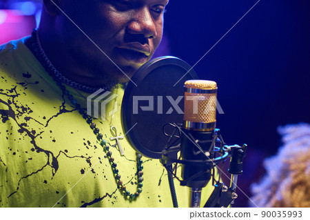 Medium close-up of stylish young Black man wearing neon yellow T-shirt standing in front of microphone recording song in studio 90035993