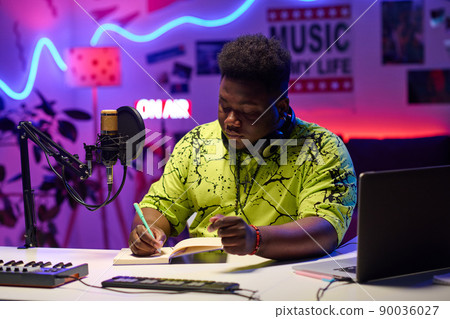 Portrait of creative young gen Z African American man sitting at desk in home studio working on broadcast or podcast text Portrait of creative young gen Z African American man sitting at desk in home studio working on broadcast or podcast text 90036027