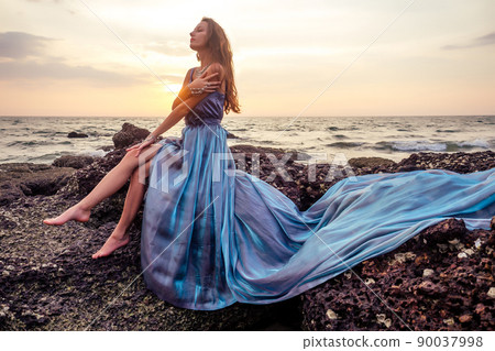 gorgeous style brunette young woman in blue chameleon dress long train standing on a rock near the sea, sand tropical on a rock.beautiful mermaid lonely melancholy waiting freedom and freelancing 90037998