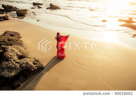 Young women wearing a red saree on the beach goa India.girl in traditional indian sari on the shore of a paradise island among the rocks and sand enjoying the freedom and the sunset 90038002