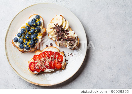 Fruity vegetarian toasts for breakfast on gray background. Bread slices with ricotta, berries, banana, chocolate and seeds. Fruity vegetarian toasts for breakfast on gray background. Bread slices with ricotta, berries, banana, chocolate and seeds. 90039265