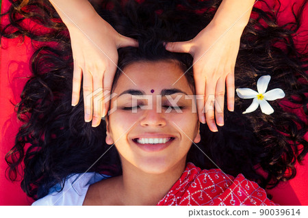 Female beautician doctor with patient make procedure to beautiful indian girl.close up portrait of asian woman closed eyes and white smile wellness head massaging on beach.Face lifting massage in spa 90039614