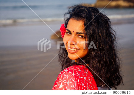 Indian female fashion model white-toothed smile with flower in her curly hair in traditional india outfit red wedding sari posing near tropical trees on a paradise beach sea ocean.wellness spa resort 90039615