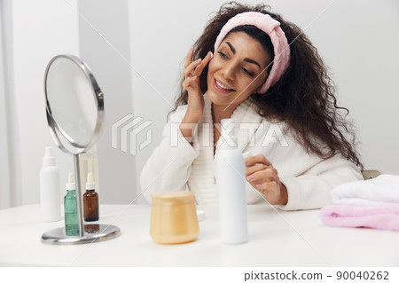 Close up portrait of enjoyed tanned pretty curly Latin lady admiring herself in mirror use facial cream in white home interior. Copy space. MIRROR, MIRROR, ON THE WALL, WHO IS THE FAIREST OF THEM ALL 90040262