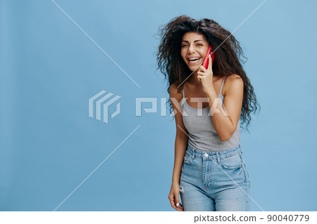 ONLINE CALL AD CONCEPT. Laughing beautiful curly Latin lady hold pink smartphone in hand, talk about holidays, look at camera. Cool offer. Studio shoot over blue sky background. Copy space for text ONLINE CALL AD CONCEPT. Laughing beautiful curly Latin lady hold pink smartphone in hand, talk about holidays, look at camera. Cool offer. Studio shoot over blue sky background. Copy space for text 90040779