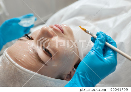 Beautiful brunette woman enjoying applying cosmetic mask with closed eyes in sunny spa center. Relaxing treatment and medicine concept. Beautiful brunette woman enjoying applying cosmetic mask with closed eyes in sunny spa center. Relaxing treatment and medicine concept. 90041799