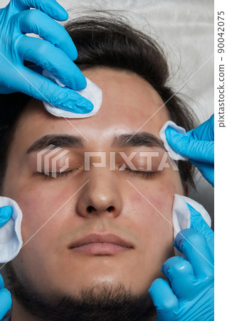 Unrecognizable cosmetologist in medical gloves removing blackheads from middle aged man nose, using tools and cotton pad, side view. Closeup of handsome man attending cosmetologist. 90042075