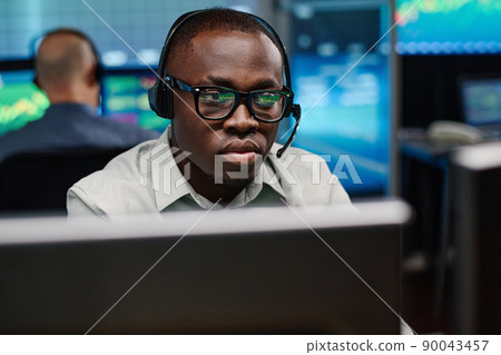 Serious young African American man wearing eyeglasses working in stock trading company watching charts and graphs of currency on computer screen 90043457