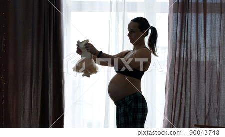 Silhouette of Pregnant Woman Examines Cute Baby Dress over Big Window at Home. Motherhood, Preparation and Expectation of Baby Concept 90043748