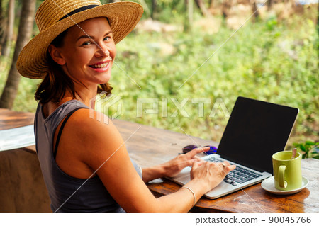 Technology and travel. Working outdoors. Freelance concept. Pretty young woman in hat using laptop in cafe on tropical beach.Hooray victory success and successful deal concept promotion at work Technology and travel. Working outdoors. Freelance concept. Pretty young woman in hat using laptop in cafe on tropical beach.Hooray victory success and successful deal concept promotion at work 90045766