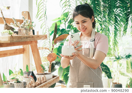 Smiling young woman taking smartphone picture of plant in a small shop Smiling young woman taking smartphone picture of plant in a small shop 90048290
