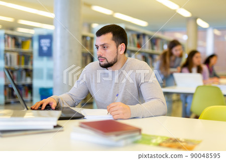 Focused young student studying with books and laptop in library Focused young student studying with books and laptop in library 90048595