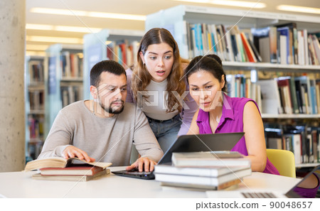 Three adult students work on laptop and read books in a public library Three adult students work on laptop and read books in a public library 90048657