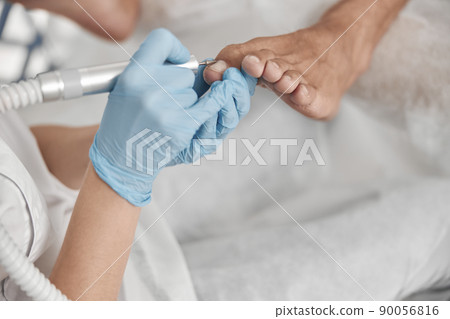 Perdicure master polishes toenail of client with grinding tool at procedure in salon 90056816