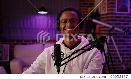 Portrait of man content creator smiling confident at camera while answering questions from listeners in home recording studio. African american online radio host reading fan mail from laptop computer. 90058157