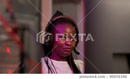 Handheld shot of african american gamer girl with professional pc setup using gaming headset talking to team members. Woman looking at computer screen while playing multiplayer online game. 90058166