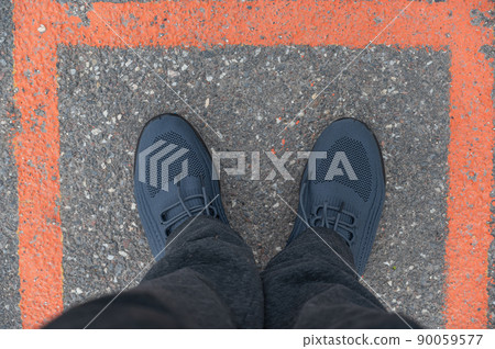 A view of my feet standing inside the orange square. A mature man in gray pants and blue sneakers standing on the pavement. Outdoors. Daytime. Selective focus. A view of my feet standing inside the orange square. A mature man in gray pants and blue sneakers standing on the pavement. Outdoors. Daytime. Selective focus. 90059577