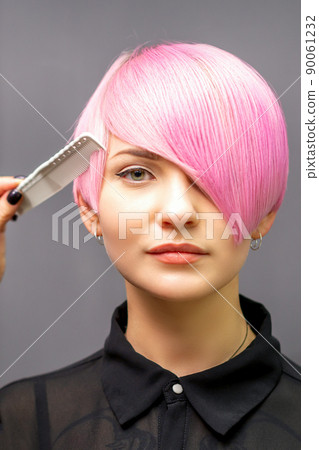 The female hairdresser is combing the dyed pink short hair of the young woman against the dark wall The female hairdresser is combing the dyed pink short hair of the young woman against the dark wall 90061232