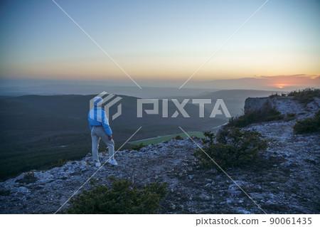 Woman tourist on top of sunrise mountain. The girl salutes the sun, wearing a blue jacket, white hat and white jeans. Conceptual design. Woman tourist on top of sunrise mountain. The girl salutes the sun, wearing a blue jacket, white hat and white jeans. Conceptual design. 90061435