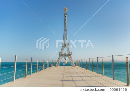 Large model of the Eiffel Tower on the beach. A woman walks along the pier towards the tower, wearing a blue jacket and white jeans. 90061436