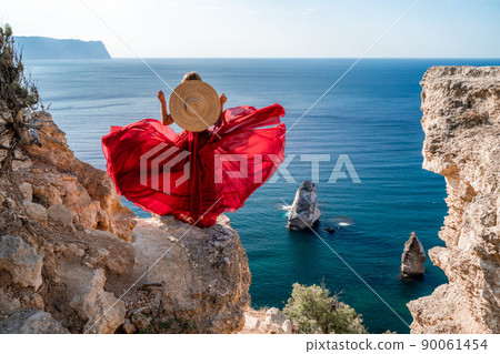 A woman in a flying red dress fluttering in the wind and a straw hat against the backdrop of the sea. A woman in a flying red dress fluttering in the wind and a straw hat against the backdrop of the sea. 90061454