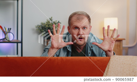 Man playing hide and seek peekaboo game hiding behind sofa looking at camera, positive smiling Man playing hide and seek peekaboo game hiding behind sofa looking at camera, positive smiling 90065987