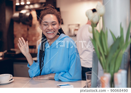 Pretty mixed-race young female bartender with piercing smiling at counter and waving hand. Pretty mixed-race young female bartender with piercing smiling at counter and waving hand. 90068490