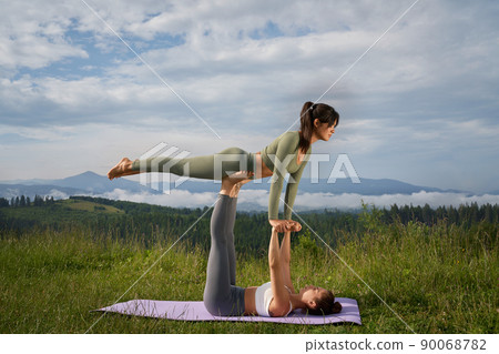 Sporty women in activewear doing acro yoga among beautiful summer mountains. Two caucasian ladies training regularly together on fresh air.  90068782