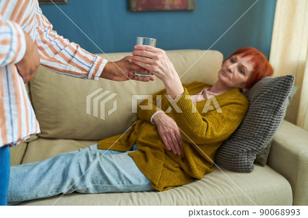 Young caregiver giving glass of water to senior woman while she lying on sofa in living room Young caregiver giving glass of water to senior woman while she lying on sofa in living room 90068993