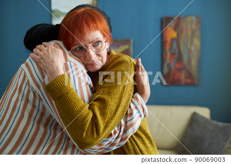 Elderly woman with red hair in eyeglasses hugging female caregiver while they standing in room 90069003