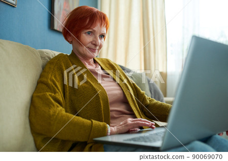 Retired woman with red hair sitting on sofa in living room and typing on laptop 90069070
