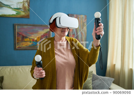 Mature woman in goggles holding joysticks in her hands and gesturing during virtual reality game at home Mature woman in goggles holding joysticks in her hands and gesturing during virtual reality game at home 90069098
