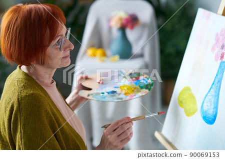 Senior red haired woman in eyeglasses looking at her picture on canvas during painting at class 90069153