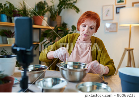 Mature woman stirring soil in bowl and telling about planting to camera on mobile phone, she streaming at home Mature woman stirring soil in bowl and telling about planting to camera on mobile phone, she streaming at home 90069235